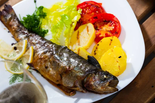 Baked whole trout with potatoes, tomatoes and greens on plate, glass of wine — Stock Photo, Image