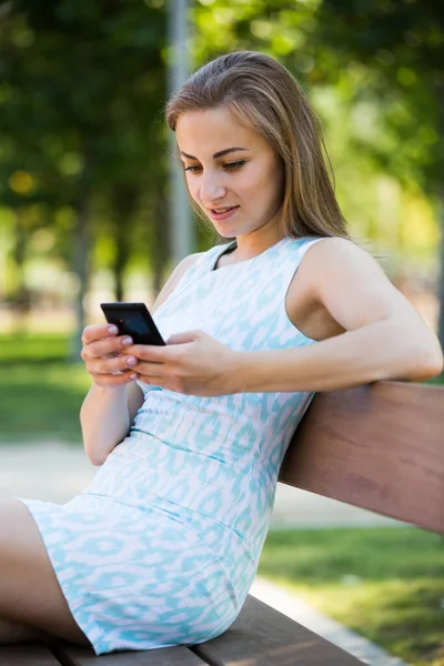 Девушка сидит на скамейке и пользуется телефоном в парке — стоковое фото