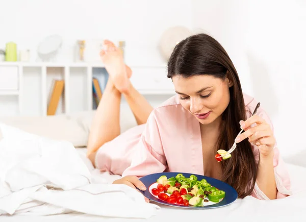 Pretty woman in bathrobe eating vegetable salad from plato in bed — Stock Photo, Image