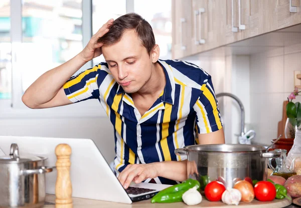 Νεαρός διαβάζει μια συνταγή για πιάτα σε ένα φορητό υπολογιστή — Φωτογραφία Αρχείου