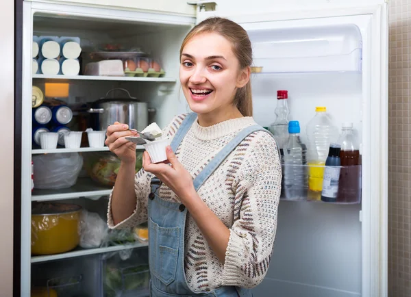 Girl eating yogurt near fridge — Stock Photo, Image