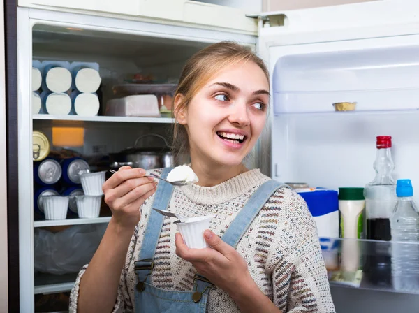 Girl eating yogurt near fridge — Stock Photo, Image