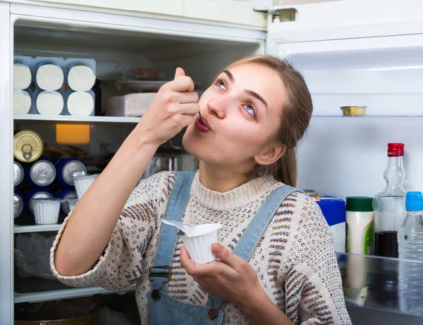 Girl eating yogurt near fridge — Stock Photo, Image