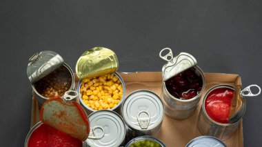 Fasulye, bezelye, mısır, karton kutuda domates gibi konserve sebzeler. Teneke kutular. Karanlık arka plan. Üst görünüm, kopyalama alanı, panorama bannesi
