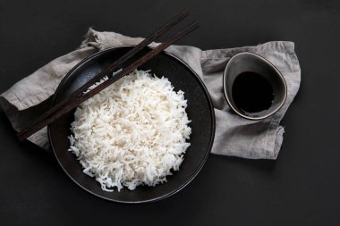 Yanında soya sosu ve yemek çubuğu olan siyah kâsede kaynamış Asya usulü pirinç. Karanlık arka plan. Üst görünüm