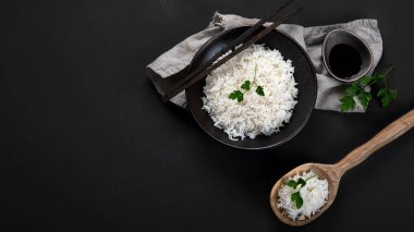 Yanında soya sosu ve yemek çubuğu olan siyah kâsede kaynamış Asya usulü pirinç. Karanlık arka plan. Üst görünüm, kopyalama alanı, panorama pankartı