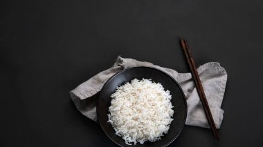 Yanında soya sosu ve yemek çubuğu olan siyah kâsede kaynamış Asya usulü pirinç. Karanlık arka plan. Üst görünüm, kopyalama alanı, panorama pankartı