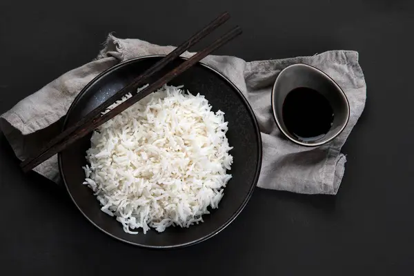 Yanında soya sosu ve yemek çubuğu olan siyah kâsede kaynamış Asya usulü pirinç. Karanlık arka plan. Üst görünüm