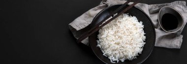 Yanında soya sosu ve yemek çubuğu olan siyah kâsede kaynamış Asya usulü pirinç. Karanlık arka plan. Üst görünüm, kopyalama alanı, panorama pankartı