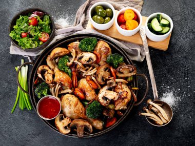 Mantar, havuç ve brokoli ile pişirilmiş tavuk butları koyu bir arka planda kızartma tavasında. Üst görünüm.