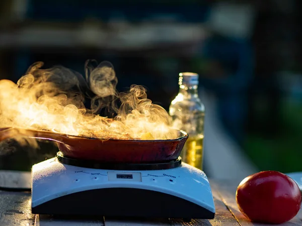 Induction cooking in the garden in the evening for dinner. Steam in the rays of the setting sun rises over a cast-iron pan with fried potatoes and chicken fillet, a tomato is on the side and there is a bottle of sunflower oil