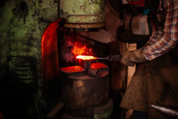 Herrero en guantes de seguridad poniendo metal calentado bajo la prensa mientras trabaja con la máquina de prensa metalúrgica. — Foto de Stock