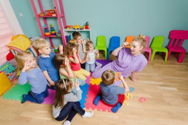 Anaokulu öğretmeni bir grup çocuğa ders veriyor, yerde oturuyor.