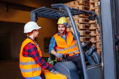 Endüstriyel forkliftte oturan erkek işçi kadın iş arkadaşıyla konuşuyor.