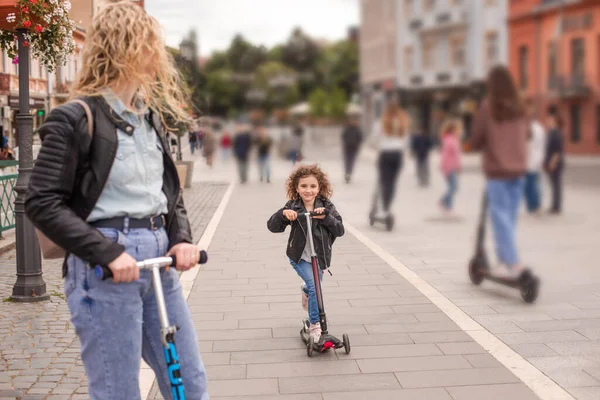 A mãe e a filha felizes estão de pé com scooters na cidade — Fotografia de Stock