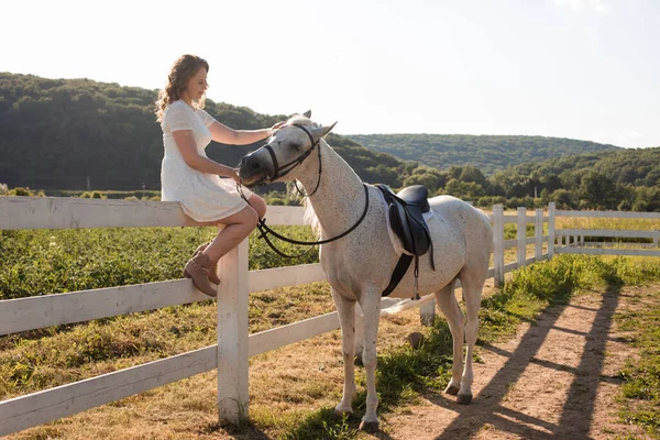 Die glückliche Frau streichelt ein Pferd, während sie auf einem Zaun sitzt — Stockfoto