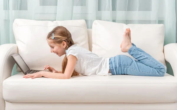 Niña acostada en el sofá mira el cuaderno y se ríe —  Fotos de Stock