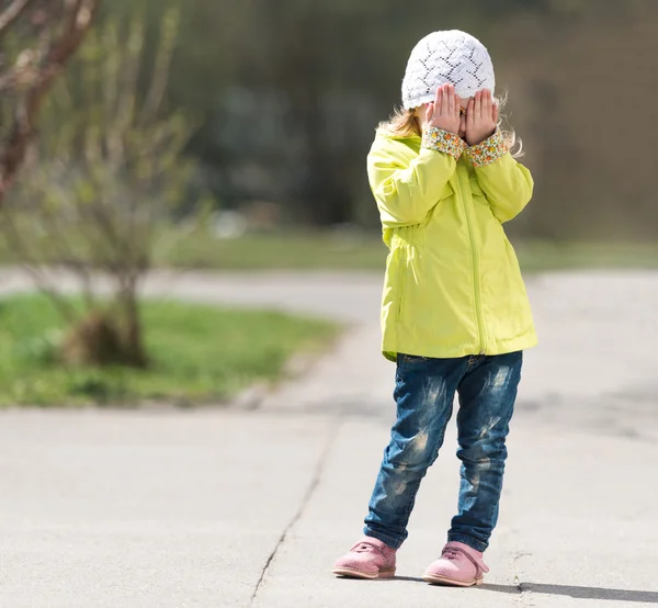 Lovely little girl in yellow coat hiding face with hands — Stock Photo, Image