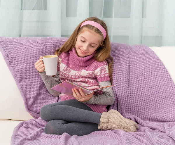 Little cute girl on sofa with cup watching a magazine — Stok fotoğraf