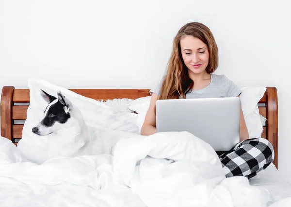 Dizüstü bilgisayarı olan ve yatakta köpeği olan kız. — Stok fotoğraf