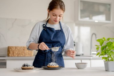 Ev Mutfağında Yaban mersinli böreğin üzerine pudra şekeri serpiştiren kız.