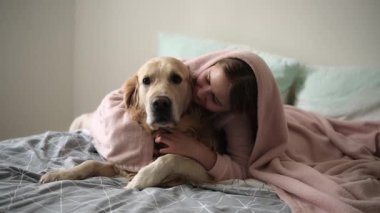 Tatlı küçük kız, köpekle yatakta oynuyor, altın av köpeğiyle battaniyeye sarılıp mutlu mesut sarılıyor.