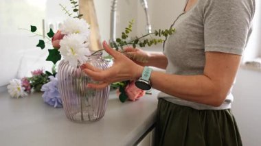 Kadın mutfaktaki vazoya yapay çiçekler koyuyor.