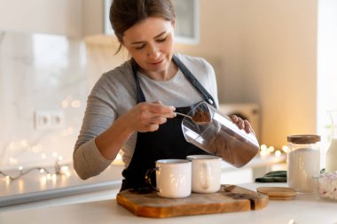 Bir kadın Noel süslemeli mutfakta lokum şekeriyle sıcak kakao hazırlıyor.