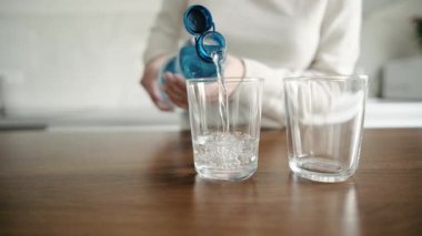 Kız mutfak masasındaki şişeden iki bardak köpüklü su dolduruyor.