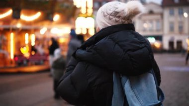 Kız, şehir meydanında yeni yıl ışıklarıyla dönen parlak bir atlıkarıncaya karşı Noel pazarında yürüyor.