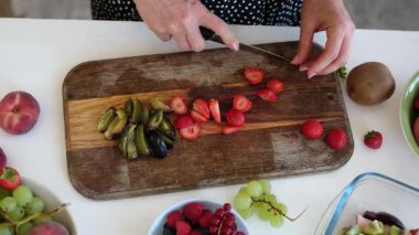 Kadın, parlak bir mutfakta tahta bir tahtanın üzerinde farklı meyveler kesiyor.