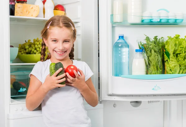 Liten flicka håller grönsaker nära kylskåp — Stockfoto