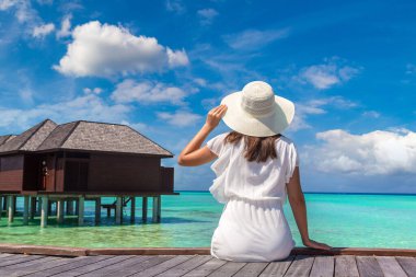 Maldivler adasındaki tropikal sahil iskelesinde (ahşap iskelede) su lüks villalarının önünde oturan güzel genç bir kadın.