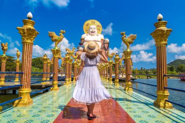Wat Plai Laem Tapınağı, Samui, Tayland 'da gülümseyen ve mutlu Buda heykelinde mavi elbise ve hasır şapka giyen kadın gezgin.