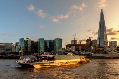 LONDON, İngiltere - 20 AĞUSTOS 2024: Thames Clippers 'ın Uber Boat' ı Thames Nehri 'ndeki Belfast savaş gemisini geçerken Londra, İngiltere, İngiltere' de altın günbatımında Çömlek gökdeleniyle