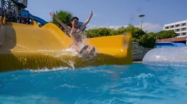 Tatil parkındaki su kaydırağından indim. Su kaydırağı aile tatilinde yavaş çekim, bikinili bir kadın mavi su damlalarından oluşan bir havuza iniyor..