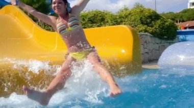 Tatil parkındaki su kaydırağından indim. Su kaydırağı aile tatilinde yavaş çekim, bikinili bir kadın mavi su damlalarından oluşan bir havuza iniyor..