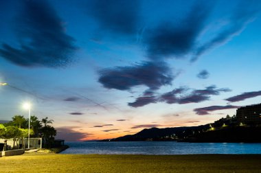 Dramatik bulutları, yumuşak akşam ışığı ve sakin deniz sularıyla sakin bir sahil manzarası. Ufuktaki sıcak tonlar derin mavi gökyüzü ile zıtlık yaratarak, barışçıl bir kıyı atmosferi yaratıyor. Seyahat, tatil, turizm için mükemmel bir geçmiş.