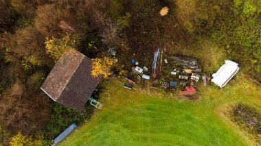 Dağınık aletler, eski makineler ve aşırı gelişmiş bitki örtüsüyle çevrili terk edilmiş bir kır evinin insansız hava aracı görüntüsü. Kırsal bozulma, terk edilmiş binalar, çevresel sorunlar, unutulmuş yerler ve kırsal yaşam gibi konular için kullanışlı.