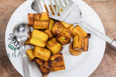 Altın kızartılmış muz dilimleri, beyaz dekoratif bir tabakta, çatal ve kaşıkla, otantik Afrika mutfağı ve geleneksel yemek pişirme yöntemleri sergileniyor.