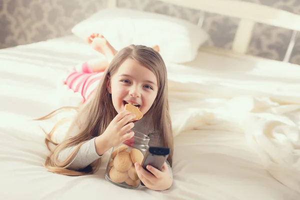 Meisje het eten van een havermout cookie en schakelt de kanalen op de TV liggend in bed — Stockfoto