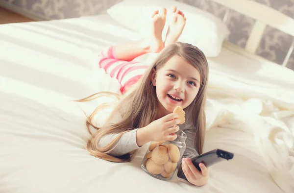 Fille manger un cookie d'avoine et échange les chaînes sur la télévision couché dans le lit — Photo