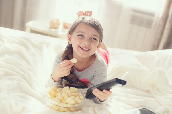 A menina alegre come pipocas e modifica canais da TV que jaz em uma cama — Fotografia de Stock