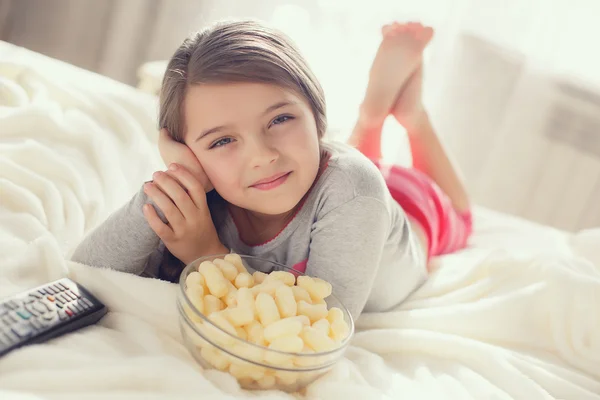 Het vrolijke meisje eet popcorn en verandert de kanalen van de TV die in een bed liggen — Stockfoto