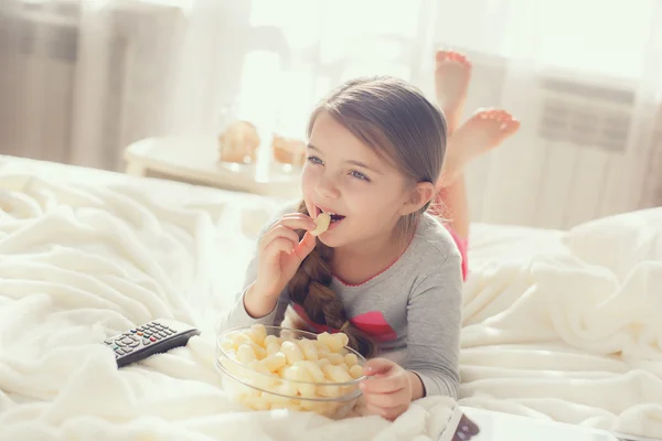 Het vrolijke meisje eet popcorn en verandert de kanalen van de TV die in een bed liggen — Stockfoto