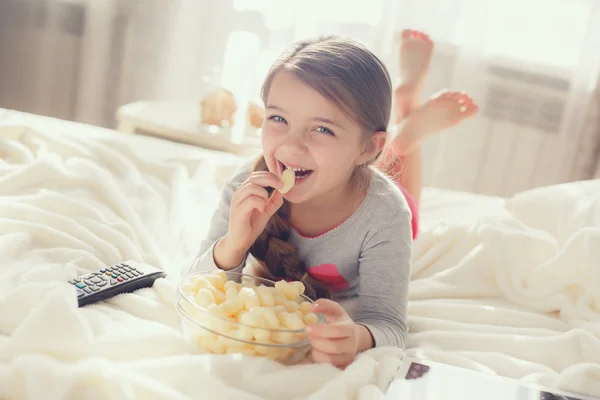 A menina alegre come pipocas e modifica canais da TV que jaz em uma cama — Fotografia de Stock