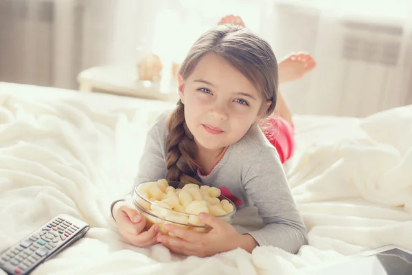 Het vrolijke meisje eet popcorn en verandert de kanalen van de TV die in een bed liggen — Stockfoto