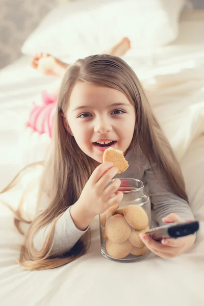 オートミールクッキーを食べて、ベッドに横たわっているテレビのチャンネルを切り替える女の子 — ストック写真