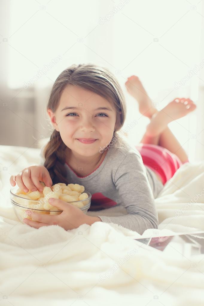 The cheerful girl eats popcorn and changes channels of the TV lying in a bed