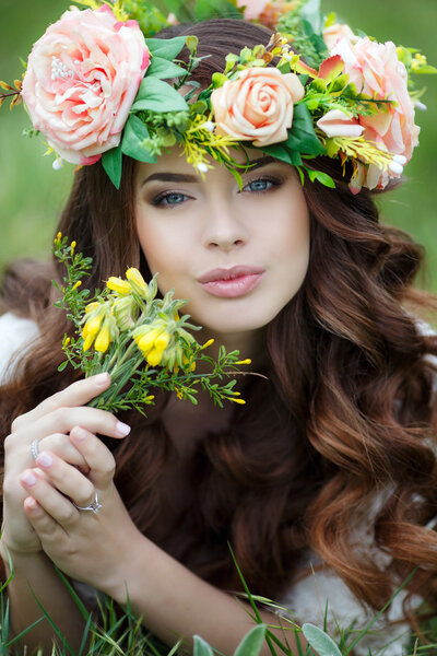 Spring portrait of a beautiful woman in a wreath of flowers,long curly red hair,gray eyes,light makeup and a beautiful smile,dressed in a white summer dress,lying on the soft green grass outdoors in spring